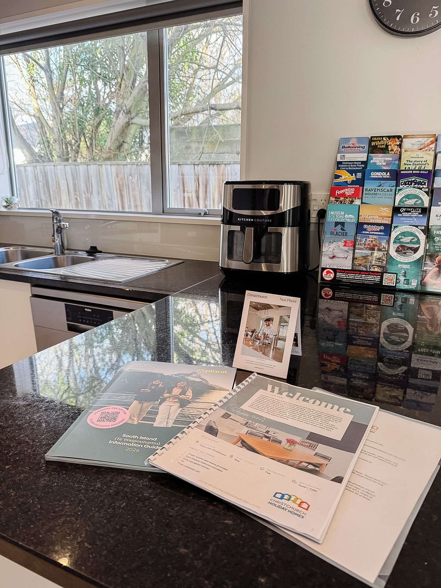 Een keukenhoek met een koffieapparaat, een raam met uitzicht op bomen, en een presentatie van reisbrochures.
