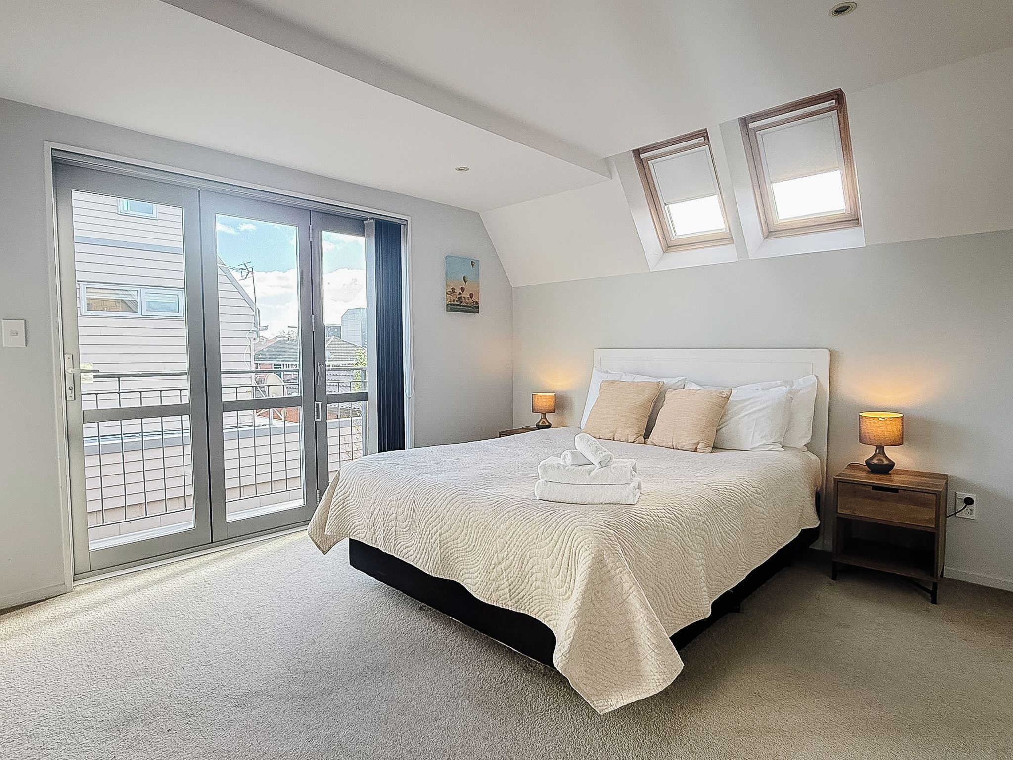 A bedroom with a bed, two bedside lamps, a sliding glass door, and a small painting on the wall.