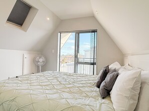 A bedroom with a large bed, a standing fan, and a window with a view of buildings.