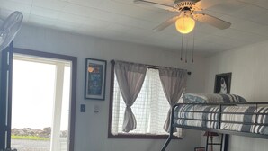 Quarto com beliche, escrivaninha com cadeira, ventoinha no teto e janela com cortinas.