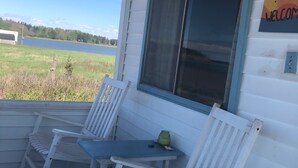 Varanda com cadeiras de balanço brancas e uma mesa, com vista para um lago e vegetação.