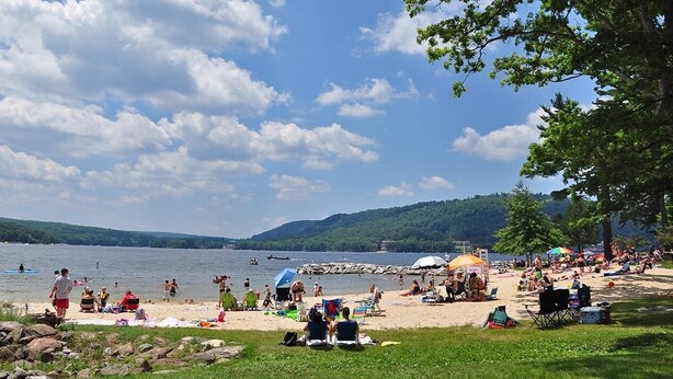A lakeside beach with people swimming and sunbathing, surrounded by greenery and hills. - Lakefront Log Home w/ Dock Slip, 30% Weekly Discount, 2 Miles from Nat. Park (Swanton)