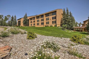 A multi-story building with a flat roof and large windows, surrounded by a landscaped area with grass and flowering plants. - Brian Head Condo w/ Pool Access - Steps to Resort! (Brian Head)