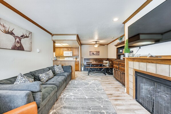 A modern living room with a gray sectional sofa, a fireplace, and a kitchen in the background. - Brian Head Condo w/ Pool Access - Steps to Resort! (Brian Head)