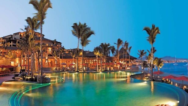 Un resort con una amplia piscina infinita, palmeras y vista al océano al anochecer.