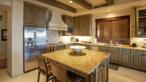 Cocina con isla central, refrigerador de acero inoxidable, armarios de madera y un amplio ventanal con persianas.