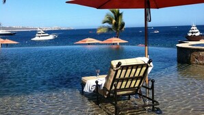 Un área junto a la piscina con una tumbona y una sombrilla, agua cristalina y barcos al fondo.