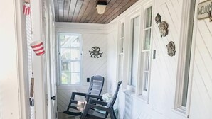 Un pasillo estrecho con paredes blancas y piso de madera, que conduce a una habitación con dos mecedoras y un ventanal.