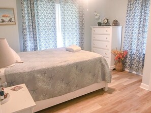 A bedroom with a bed, a dresser, a nightstand, and a window with patterned curtains. - Cute Bungalow, Hot tub, game room,fenced yard (Sneads Ferry)