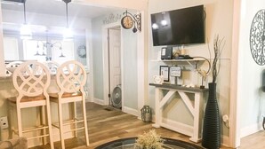 Una sala de estar con una mesa de centro redonda de vidrio, dos sillas de madera, un televisor montado en la pared y un reloj decorativo.