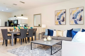 A modern living room with a grey sofa, a coffee table, and a dining area with chairs and a table. There are abstract paintings on the wall and a kitchen in the background.