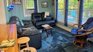 Una sala de estar acogedora con un sofá de cuero, una mesa de centro de madera y un sillón de mimbre. Hay dos ventanas con cortinas azules, una chimenea y un estante de pared con jarrones azules.