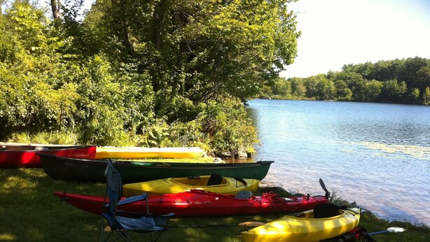 Una zona verde junto al lago con varias kayaks y una canoa, una silla plegable y un remo.