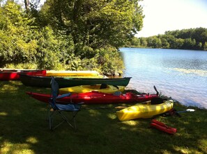 A grassy lakeside area with several kayaks and a canoe, a folding chair, and a paddle. - Fun Family Wolfeboro Winni Back Bay Waterfront with 3 SUP, 5 Kayaks & 2 Canoes!! (Wolfeboro)