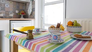Eine Küche mit einer bunten, gestreiften Tischdecke, einer Kaffeekanne, Tassen, einer Obstschale und einem Teller Kekse.