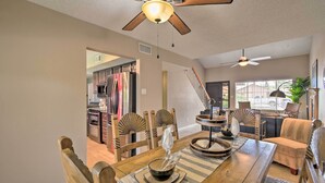 A dining area with a wooden table, chairs, and a ceiling fan. There is a kitchen visible in the background.