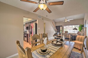 A dining area with a wooden table, chairs, and a ceiling fan. There is a kitchen visible in the background.
