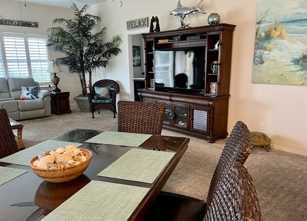 A living room with a dining table, wicker chairs, a large television, and a potted plant.