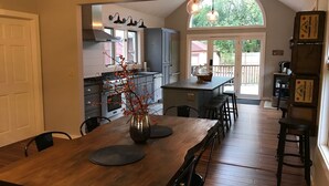 A modern kitchen with a large island, a dining table with chairs, and a view of the backyard through a glass door. - Updated Middlebury Home! Walk to Town, Sleeps Groups Comfortably (Middlebury)