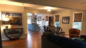 A spacious living room with a large sofa, wooden flooring, and a dining area with a kitchen in the background. - Updated Middlebury Home! Walk to Town, Sleeps Groups Comfortably (Middlebury)