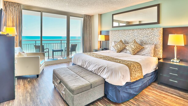 Une chambre d’hôtel spacieuse, dotée d’un grand lit, d’une table de chevet, d’une petite table, d’une chaise, d’un banc et d’un balcon offrant une vue sur la mer.