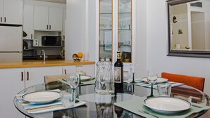 Un coin repas avec une table ronde en verre, dressée pour quatre personnes, entourée d’armoires blanches et d’une cuisine en arrière-plan.
