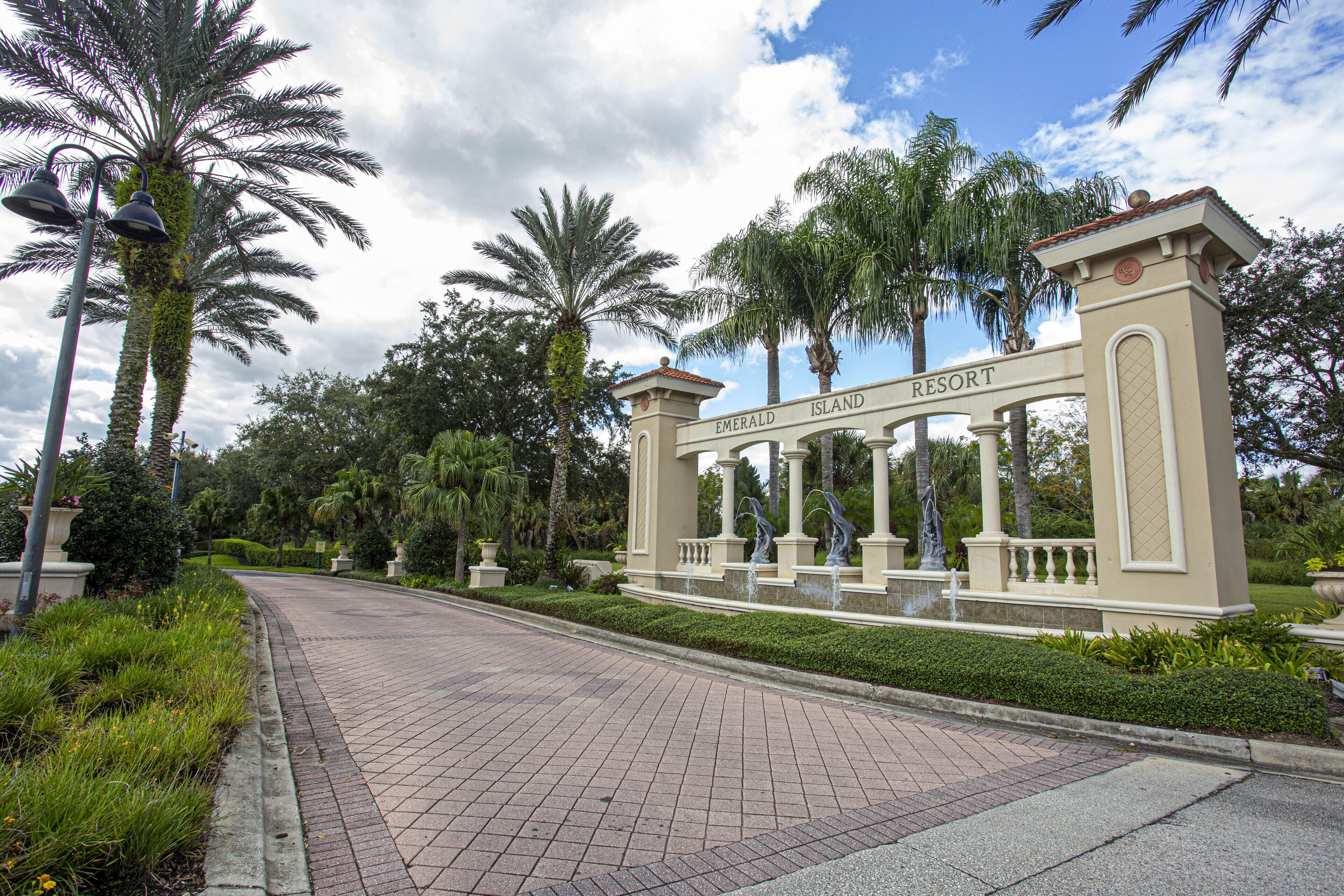 Una entrada protegida por un portón a Emerald Island Resort, con un camino pavimentado, palmeras y una fuente.