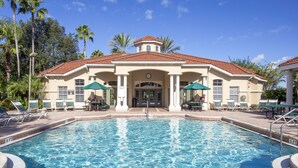 A swimming pool with a 5 FT depth, surrounded by lounge chairs and umbrellas, with a building featuring a tiled roof and palm trees in the background.