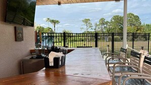 A covered outdoor bar area with a wooden ceiling, a flat-screen TV mounted on the wall, and a view of the surrounding landscape.