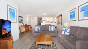 A living room with a sofa, a coffee table, and a TV. There is a kitchen in the background.