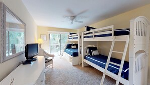 Ein Zimmer mit Stockbetten, einem Schreibtisch mit Computer, einem Deckenventilator und einem Fenster mit Blick auf Palmen.