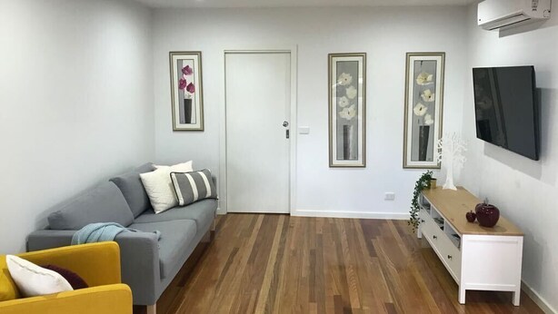 A living room with a gray sofa, a yellow armchair, a wooden floor, a white door, and a TV mounted on the wall. - Bacchus Marsh Villa unit 1 (Darley)