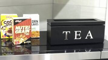 Un comptoir de cuisine avec une boîte noire étiquetée «
THÉ
» et trois boîtes de céréales : «
COCO Pops
», «
NUTRIGRAIN
» et «
CORN LAKES
».