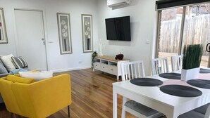 A modern living room with a yellow armchair, white dining table with black placemats, and a flat-screen TV mounted on the wall. - Bacchus Marsh Villa unit 1 (Darley)