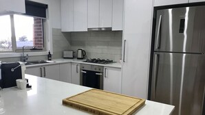 A modern kitchen with white cabinets, a stainless steel refrigerator, a wooden cutting board on the counter, and a window with a view of the outside. - Bacchus Marsh Villa unit 1 (Darley)