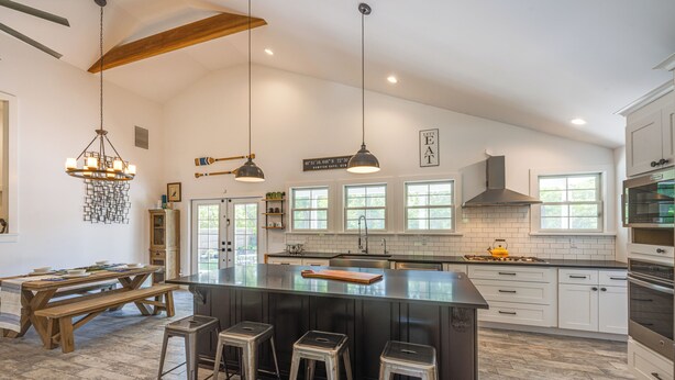 A spacious kitchen with a large island, wooden bar stools, pendant lighting, and a view of the outdoors through large windows. - Distinctive Hamptons Home w/ Pool Near Bay, Ocean (Hampton Bays)