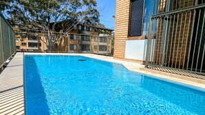 Uma piscina retangular com água azul cristalina, rodeada por um deck de madeira e uma cerca de metal.