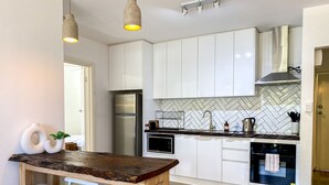 Cozinha moderna com armários brancos, painel de backsplash em padrão herringbone, geladeira de aço inoxidável e área de bar com bancada de madeira e banquetas.