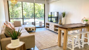Uma sala de estar moderna com sofá, mesa de centro, televisão e área de jantar com mesa de madeira e banquetas brancas.