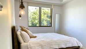 Quarto com estrutura de cama de madeira, roupa de cama branca, um banquinho de madeira redondo, janela com vista para árvores e luminária pendente.