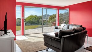 A modern living room with a red wall, a black leather sofa, a flat-screen TV, and a sliding glass door leading to an outdoor deck. - Sisters Beach Retreat (sisters beach)