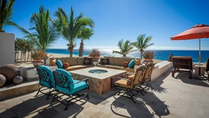 An outdoor seating area with a fire pit, sofas, and chairs, overlooking a beach with palm trees and the ocean. - 2 FREE NIGHTS IN CABO at Casa Alcini! (Cabo San Lucas)