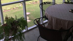 A conservatory with a view of a pond and surrounding greenery. Inside, there is a table with a floral tablecloth and a chair.
