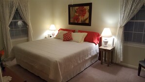 A neatly made bed with a white comforter and red and white pillows, two bedside lamps, and a framed floral painting on the wall.