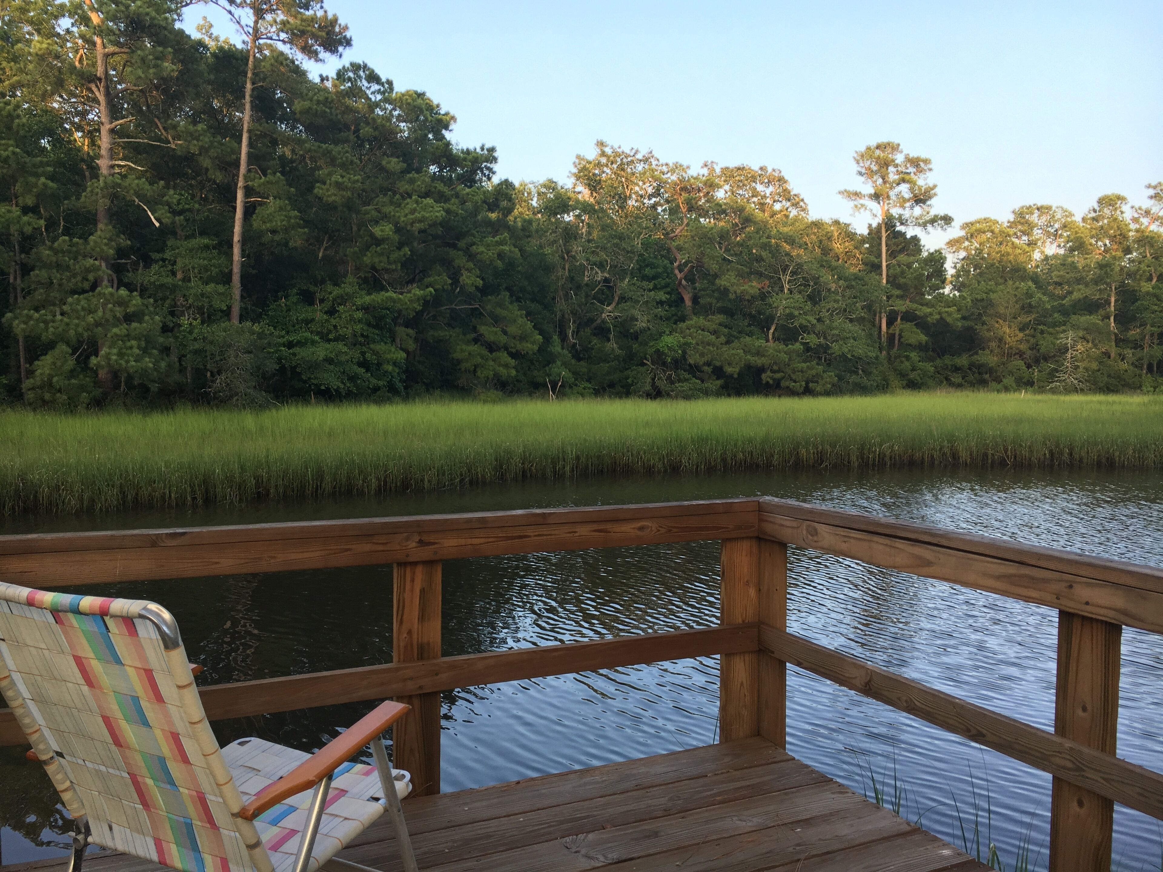 A wooden deck with a chair and table, overlooking a calm body of water and dense greenery.