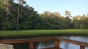 A wooden deck with a chair and table, overlooking a calm body of water and dense greenery.