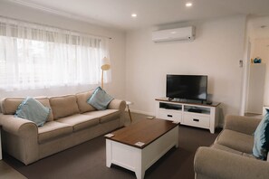 A living room with a beige sofa, a coffee table, a TV stand, and an air conditioner. - At The Beach Phillip Island - Beachley Bungalow 2 with WiFi (Surf Beach)