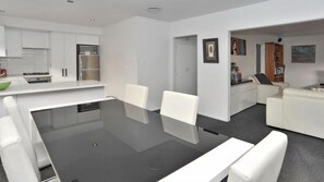 Une cuisine moderne avec des meubles blancs, un réfrigérateur et une cuisinière. On y trouve un coin repas avec une table noire et des chaises blanches. La pièce est dotée d’un tapis gris et de murs blancs.