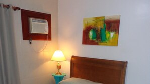 A single bed with a wooden headboard, a bedside table with a lamp, a wall-mounted heater, and a painting on the wall. - MAKE YOURSELF AT HOME! Spacious, 4 bedrooms, air conditioning, free Wi-Fi, close to the street market! (Cabo Frio)