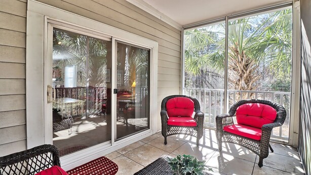 A covered patio with two wicker chairs with red cushions, a small table, and a glass door leading to an outdoor area with palm trees. - 1021 Willow Bend - Barefoot Resort Rentals (North Myrtle Beach)